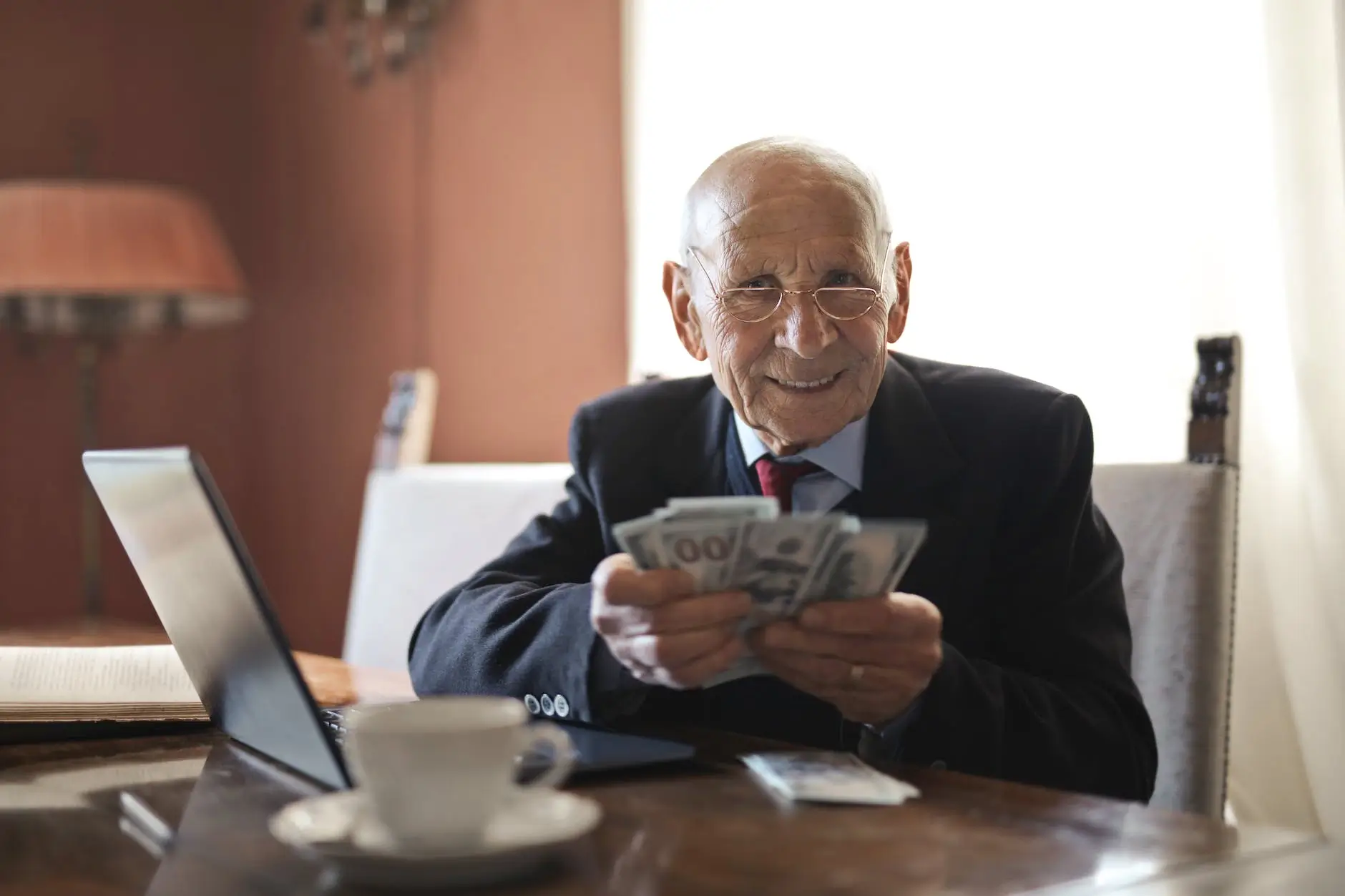 elderly businessman counting money at his desk with a laptop and coffee cup nearby
