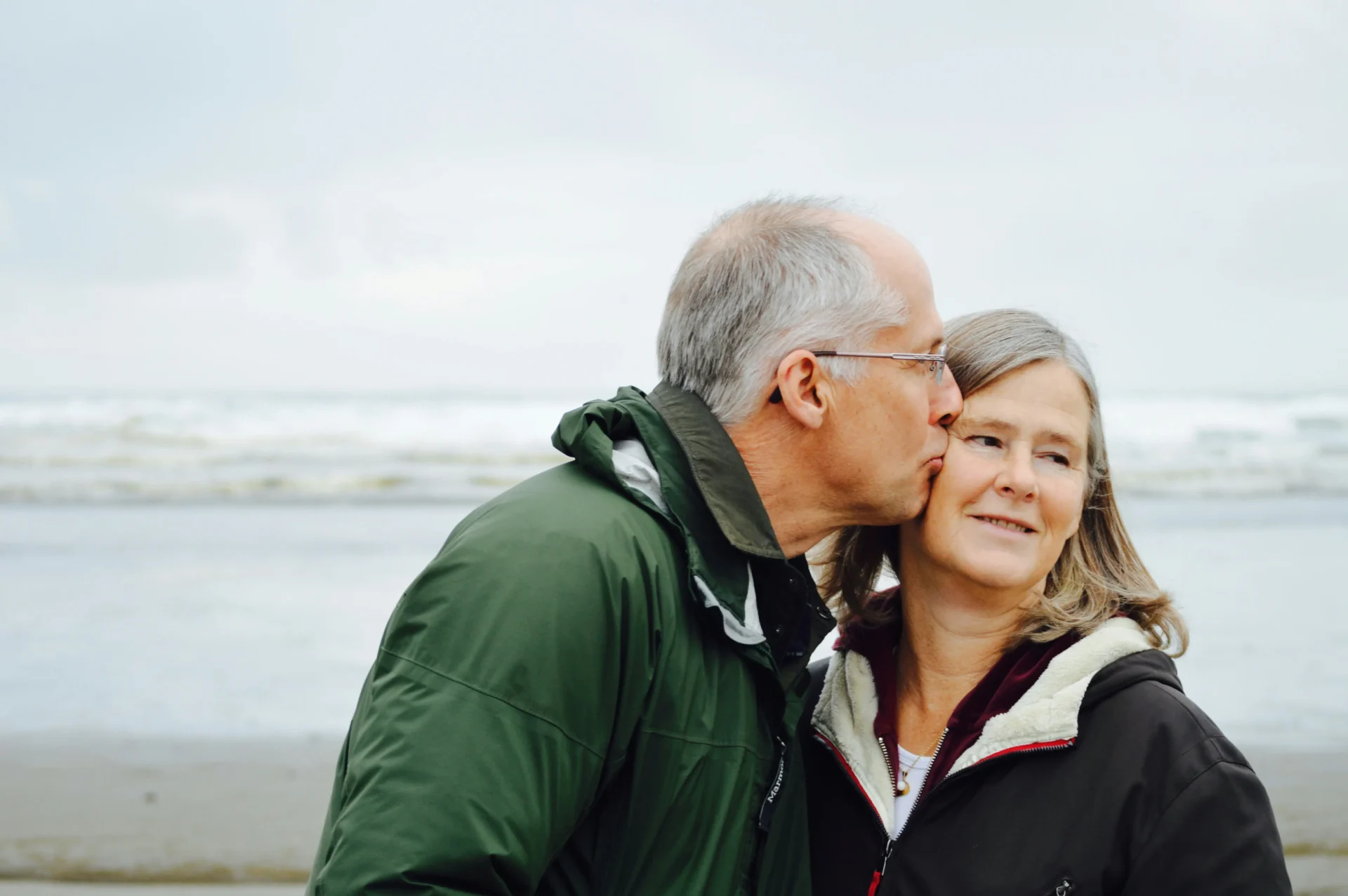 An older man kisses an older woman on the cheek while standing on a beach, both wearing jackets, sharing a moment that feels just like home.