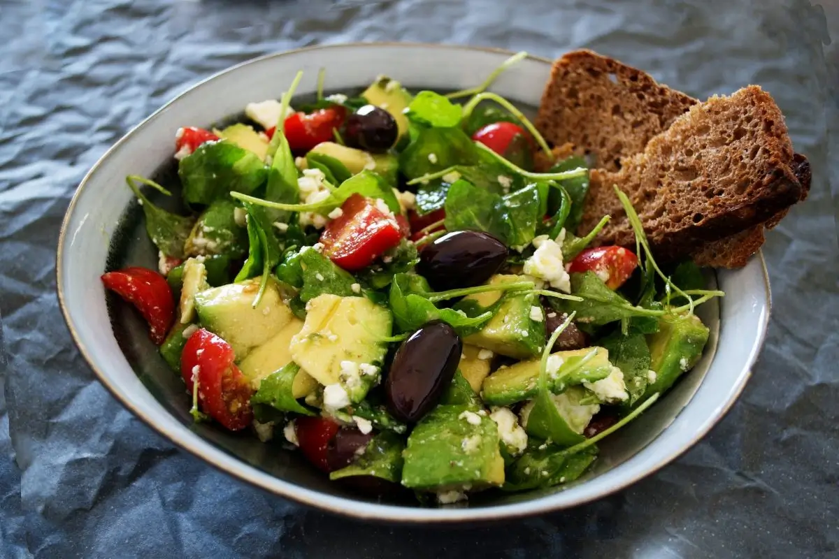 A fresh salad with spinach, avocado, strawberries, feta cheese, and black olives, served with a slice of whole grain bread in a grey bowl.