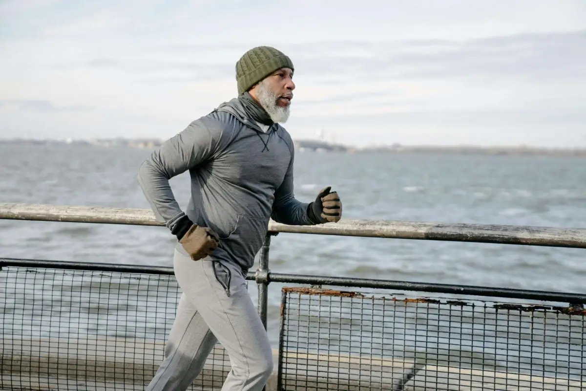 A middle-aged man jogging along a waterfront railing, wearing a gray jacket, and green beanie