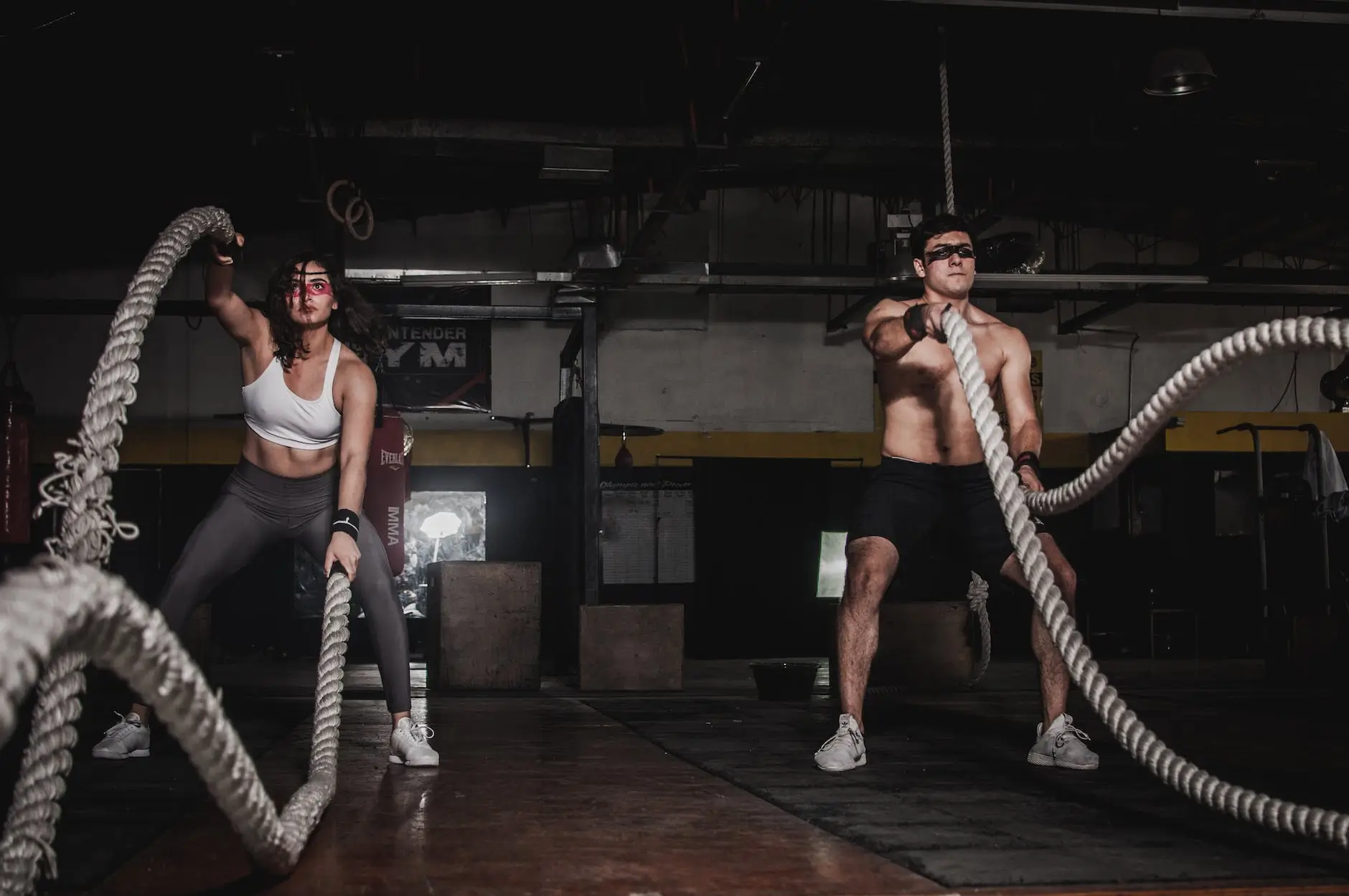 Two people, a woman and a man, exercise with battle ropes in a gym, focused and intense. brightly lit, contrast with the dim background.